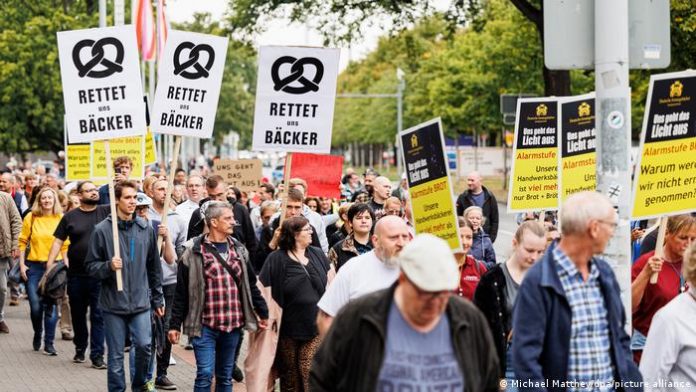 Men and women in everyday clothes walk through the city center of Hanover on a train.  Several demonstrators hold up signs reading Save us bakers. 