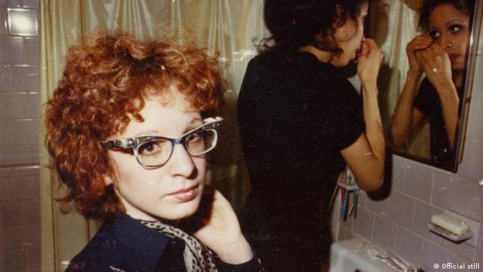 A woman with curly red hair and glasses is standing in the bathroom;  another woman is standing at the mirror in the background and doing something in her face. 
