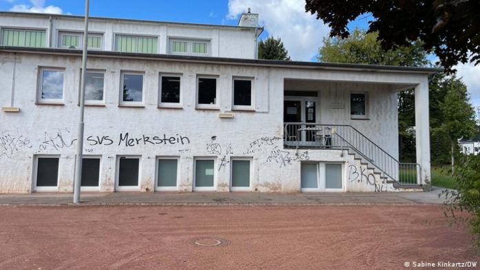 Gymnasium in Herzogenrath-Merkstein with stairs and entrance on the right.  In the front there are two rows of windows, one above the other.  The facade is white but shows clear traces of dirt and the red sand of the sports field in front and scribbles in black paint.