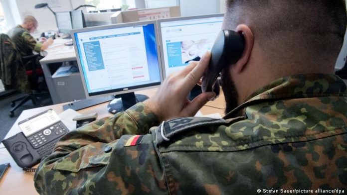 A uniformed Bundeswehr soldier makes a phone call while sitting at a desk with two computer screens. 