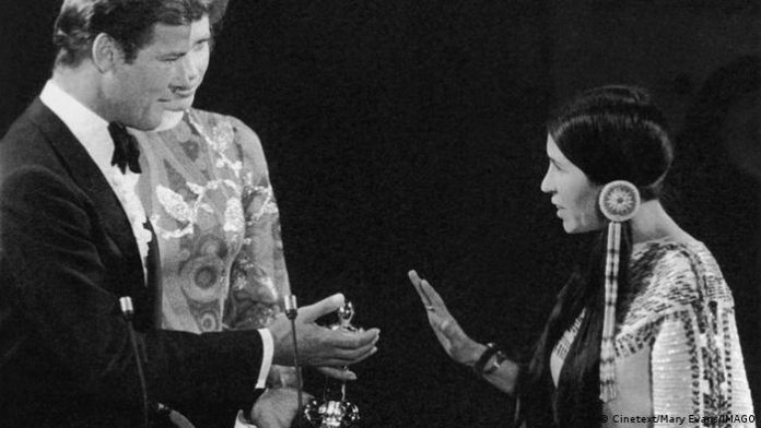Sacheen Littlefeather is dead | culture | DW Sacheen Littlefeather makes a defensive hand gesture, Roger Moore looks at her with trophy in hand, Liv Ullman in the middle